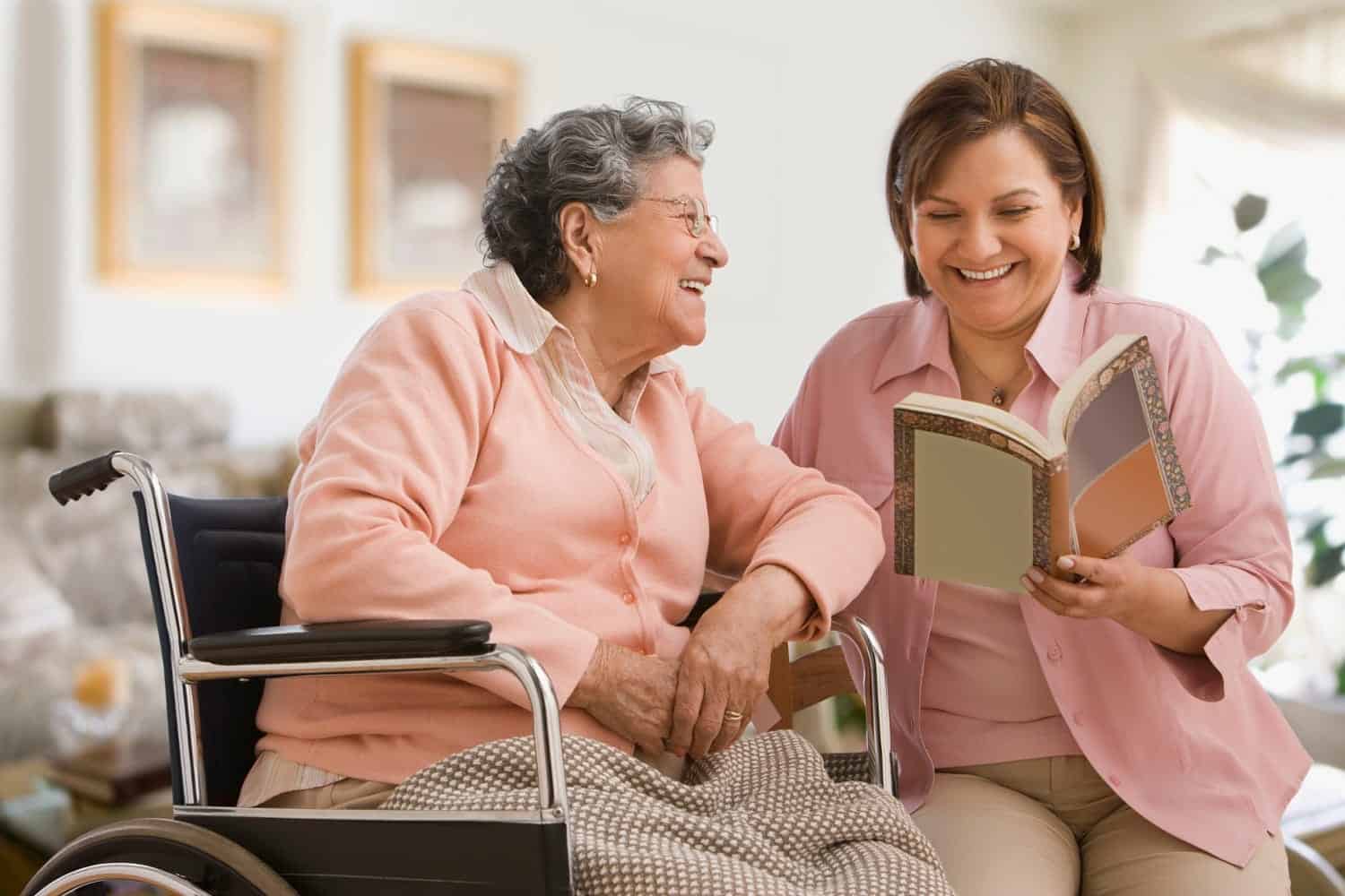 a woman in a pink shirt reading a book to an elderly woman in a wheelchair