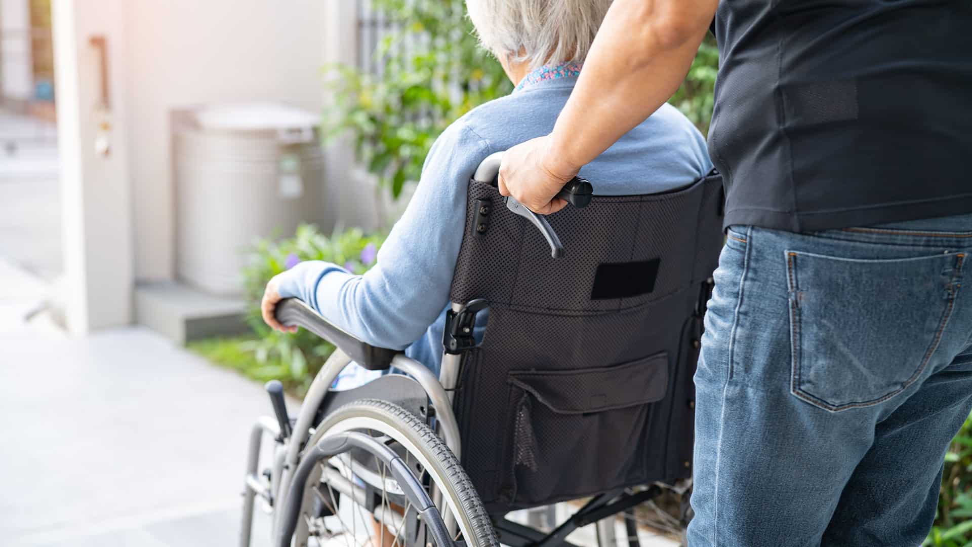 Photograph of an older adult in a wheelchair being carried by their caregiver.